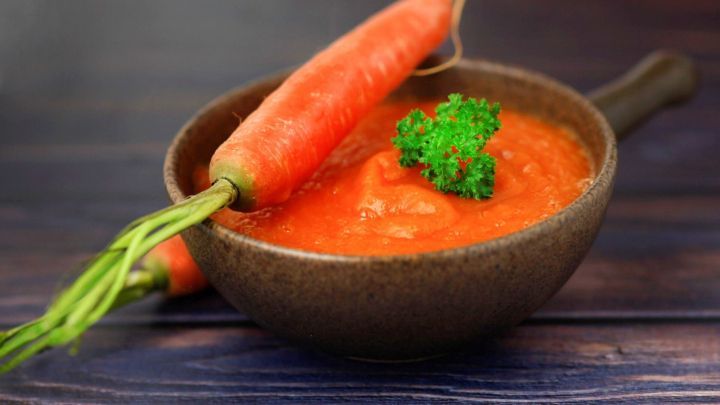 Eine Sch&uuml;ssel mit orangener Karottensuppe, garniert mit frischer Petersilie, steht auf einem Holzuntergrund.