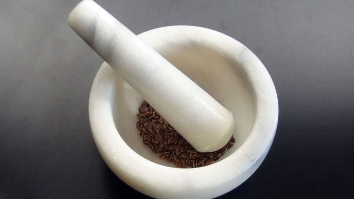 Ein M&ouml;rser aus wei&szlig;em Marmor mit einem St&ouml;&szlig;el liegt auf einer dunklen Oberfl&auml;che, w&auml;hrend Gew&uuml;rze im Inneren zersto&szlig;en werden.