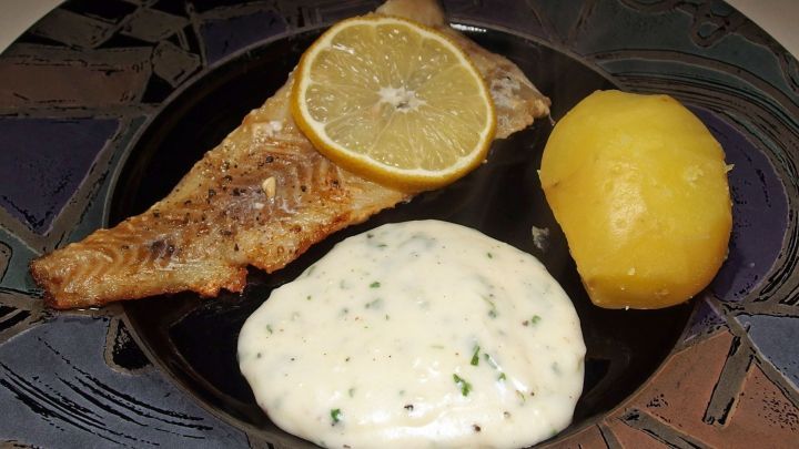Ein gebratener Fisch liegt auf einem Teller, begleitet von Petersilienso&szlig;e und einer Zitrone sowie einer gekochten Kartoffel.