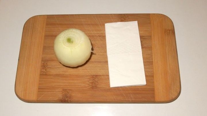 Eine halbe Zwiebel liegt auf einem Holzbrett neben einem wei&szlig;en K&uuml;chentuch, bereit zum Schneiden ohne Tr&auml;nen.