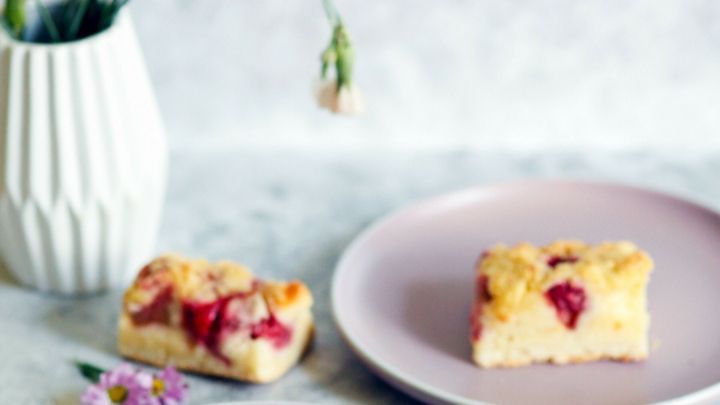 Ein geschnittener Rhabarberkuchen liegt auf Tellern, umgeben von einer Vase mit frischen Blumen auf einem hellen Tisch.