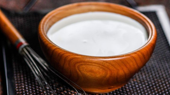 Eine Holzsch&uuml;ssel mit Rama Cremefine steht neben einem Schneebesen auf einem Gitter. Der Fokus liegt auf der cremigen Konsistenz.