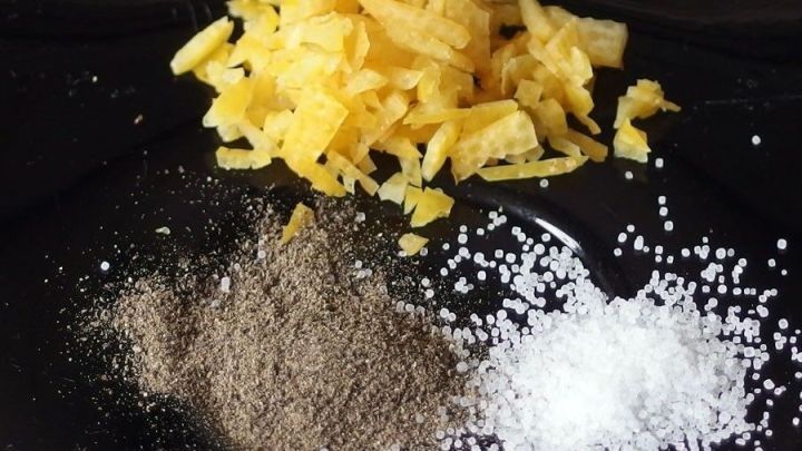 Geriebene Biozitronenschale, Salz und Pfeffer liegen auf einem schwarzen Teller und warten darauf, f&uuml;r ein Gericht verwendet zu werden.