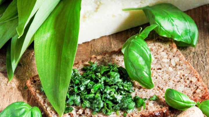 Ein trockener Roggenbrot-Schnitt mit B&auml;rlauch-Aufstrich ist auf einem Holzbrett angerichtet, umgeben von frischem Basilikum und Parmesan.