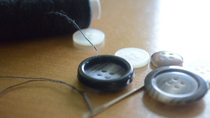 Ein schwarzer Knopf mit vier Knopfl&ouml;chern liegt auf einem Tisch, w&auml;hrend ein Faden und eine Nadel bereit zum Ann&auml;hen sind.
