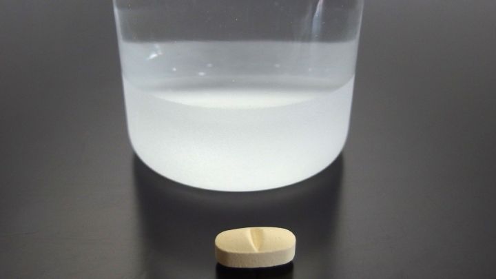 Eine Tablette liegt vor einem halbgef&uuml;llten Glas Wasser auf einer schwarzen Oberfl&auml;che, bereit zum Schlucken.