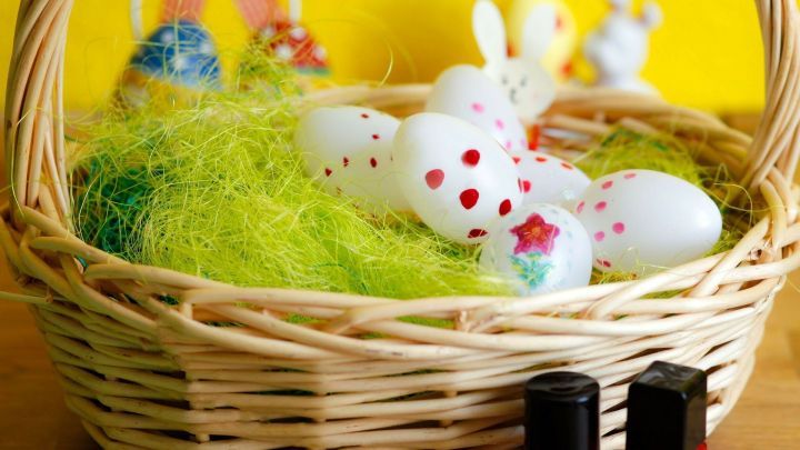 Ein Korb mit wei&szlig;en Ostereiern, die mit Nagellack bemalt sind, steht auf gr&uuml;nem Gras. Im Hintergrund sind &ouml;sterliche Dekorationen.