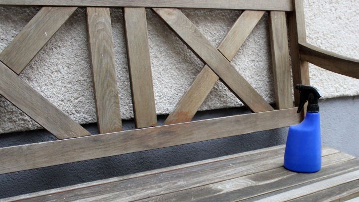 Eine blaue Spr&uuml;hflasche steht neben einer Holzbank, w&auml;hrend der Mensch die Bank mit Farbe behandelt.