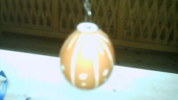 Ein h&auml;ngendes, orangefarbenes Teelicht mit einer dekorativen Musterung schwingt sanft &uuml;ber einer hellen Tischoberfl&auml;che.