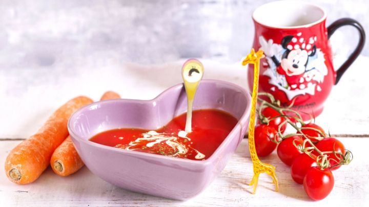 Eine herzf&ouml;rmige Sch&uuml;ssel fasst Tomatensuppe; daneben stehen Karotten und eine rote Tasse mit einem kindlichen Design.
