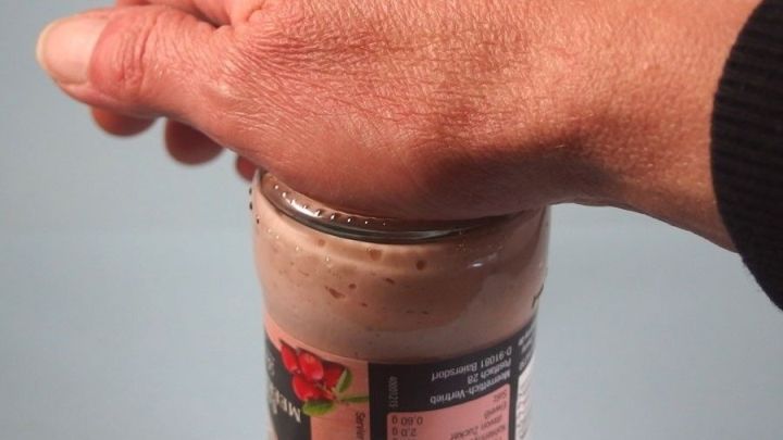 Eine Hand dr&uuml;ckt auf den Deckel eines Einmachglases, w&auml;hrend das Glas auf einem blauen Untergrund steht.