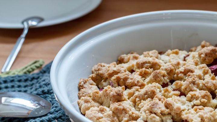 Ein Rhabarber-Crumble befindet sich in einer wei&szlig;en Sch&uuml;ssel, umgeben von einem L&ouml;ffel und einem Teller auf einem Holztisch.