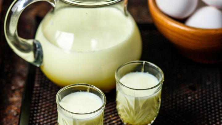 Ein Krug mit hausgemachtem Eierlik&ouml;r steht neben zwei Gl&auml;sern auf einem Tablett, w&auml;hrend im Hintergrund eine Sch&uuml;ssel mit Eiern zu sehen ist.