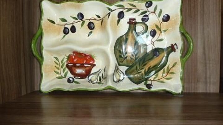 Ein dekorativer Servierteller mit Oliven- und Flaschenmotiven steht auf einem Holzregal und dient als stilvolle Stehhilfe.