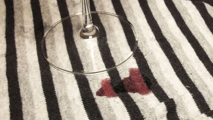 Ein Weinglas steht auf einem gestreiften Tuch, w&auml;hrend ein Rotweinfleck auf dem Stoff sichtbar ist.
