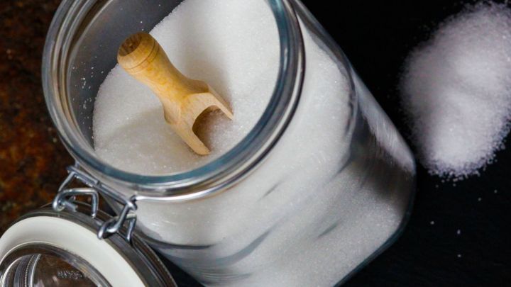 Ein Glas mit kristallinem Zucker steht offen, w&auml;hrend ein Holzl&ouml;ffel im Inneren liegt und etwas Zucker daneben verstreut ist.