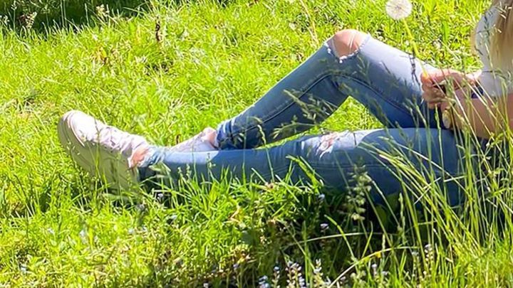 Dunkelblaue Jeans mit Rissen liegen auf saftigem, gr&uuml;nem Gras, w&auml;hrend eine Hand einen L&ouml;wenzahn h&auml;lt.