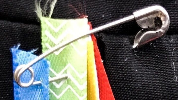 Bunte Reste von Geschenkband sind auf einer Sicherheitsnadel aufgef&auml;delt, vor schwarzem Hintergrund angespannt.