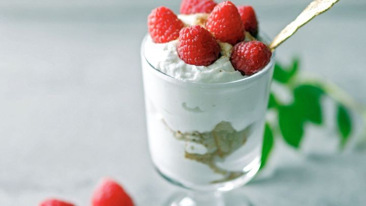 Das schichtweise Himbeer-Schichtdessert in einem Glas wird mit frischen Himbeeren garniert und steht auf einem grauen Untergrund.