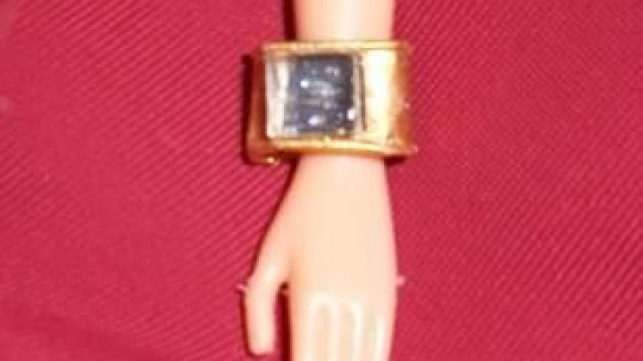 Ein goldenes Armband aus einem T&uuml;tenverschluss schm&uuml;ckt das Handgelenk einer Barbie-Puppe auf rotem Untergrund.