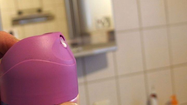 Ein Haarspray-Flakon wird in einem Badezimmer gehalten, mit Blick auf einen Spiegel und Fliesen im Hintergrund.