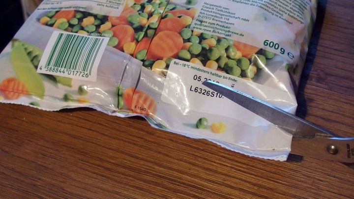 Eine T&uuml;te mit Tiefk&uuml;hlware liegt auf einem Tisch, w&auml;hrend eine Schere verwendet wird, um sie von unten aufzuschneiden.