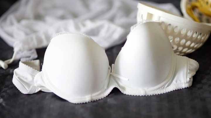 Ein wei&szlig;er B&uuml;gel-BH liegt auf einer dunklen Oberfl&auml;che, umgeben von einem W&auml;schekorb und einem W&auml;schesack, bereit f&uuml;r die Maschinenw&auml;sche.