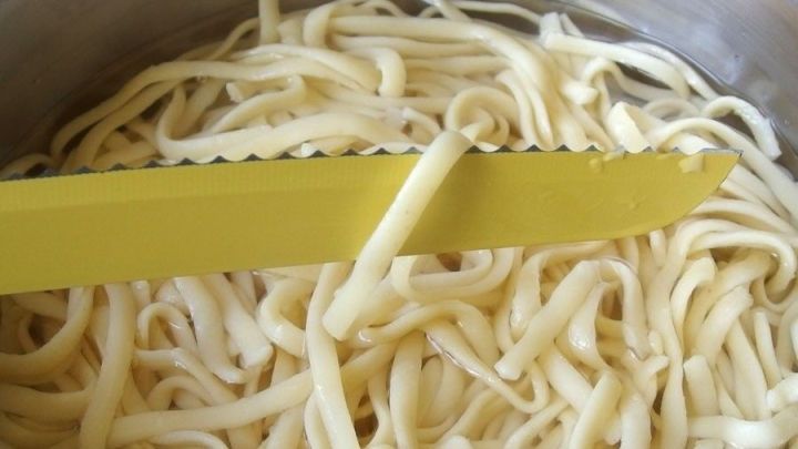 Ein einzelnes Spaghetti wird mit einem gelben Messer aus einem Topf mit kochendem Wasser gehoben, um die Garprobe zu testen.