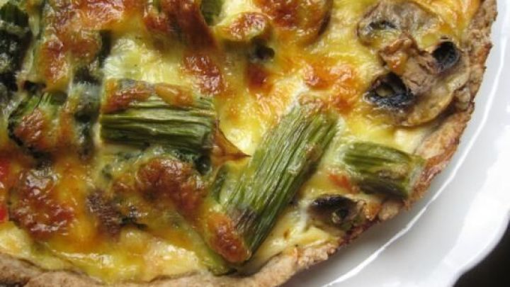 Ein k&ouml;stlicher Fr&uuml;hlingskuchen zeigt gebr&auml;unten Spargel, Brokkoli und Champignons auf einem goldgelben Teig, serviert auf einem wei&szlig;en Teller.