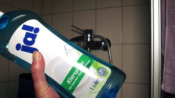 Eine Hand h&auml;lt eine Flasche Geschirrklarsp&uuml;ler vor einer Duschwand, bereit zum Reinigen.