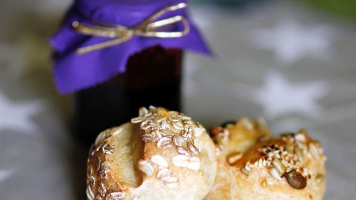 Zwei knusprige Br&ouml;tchen mit Haferflocken liegen neben einem Glas Marmelade mit lila Deckel auf einem Tisch.