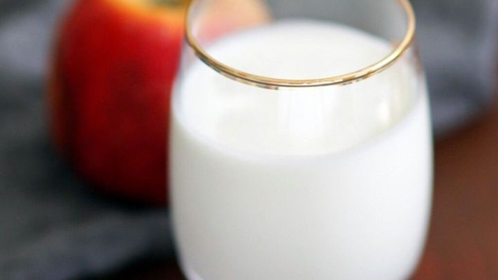 Ein Glas cremiger Joghurt-Buttermilch steht neben einem roten Apfel auf einem grauen Tuch, ideal f&uuml;r ein Fr&uuml;hst&uuml;cksgetr&auml;nk.