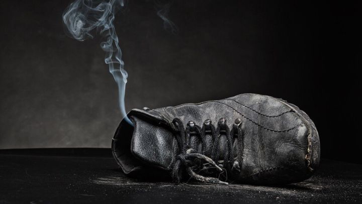 Ein abgenutzter Schuh liegt auf einem dunklen Untergrund, w&auml;hrend Rauch aus der &Ouml;ffnung aufsteigt, was stinkende Schuhe symbolisiert.