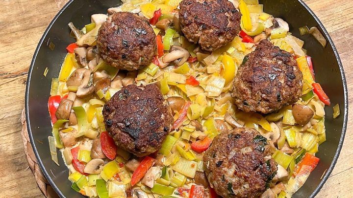 Kleine Frikadellen ruhen auf einem bunten Gem&uuml;sebett aus Lauch, Paprika und Pilzen in einer Pfanne auf Holz.