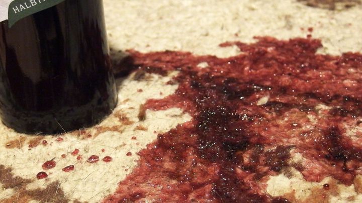Eine Weinflasche steht neben einem Rotweinfleck auf einem Teppich, der die Umgebung mit einem tiefroten Farbton verunreinigt.