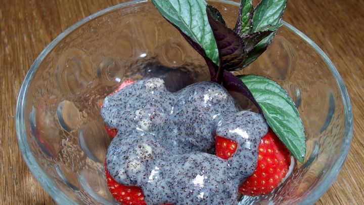 Saftige Erdbeeren liegen in einer Sch&uuml;ssel, &uuml;berzogen mit wei&szlig;er Schokolade und Mohn, umgeben von frischen Bl&auml;ttern.