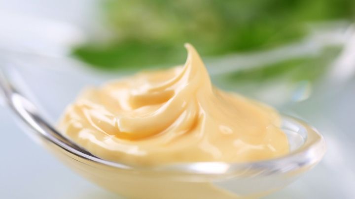 Eine cremige Mayonnaise aus gekochtem Eigelb in einem L&ouml;ffel, umgeben von frischen gr&uuml;nen Kr&auml;utern im Hintergrund.