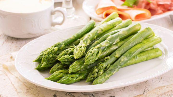 Gr&uuml;ner Spargel liegt frisch auf einem wei&szlig;en Teller, umgeben von einer Tasse und Aufschnitt auf einem rustikalen Tisch.