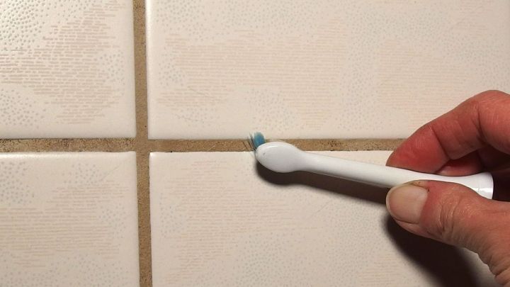 Eine Hand h&auml;lt eine Zahnb&uuml;rste, w&auml;hrend sie schwarze Fugen im Bad reinigt, um sie aufzuhellen. Der Hintergrund zeigt helle Fliesen.