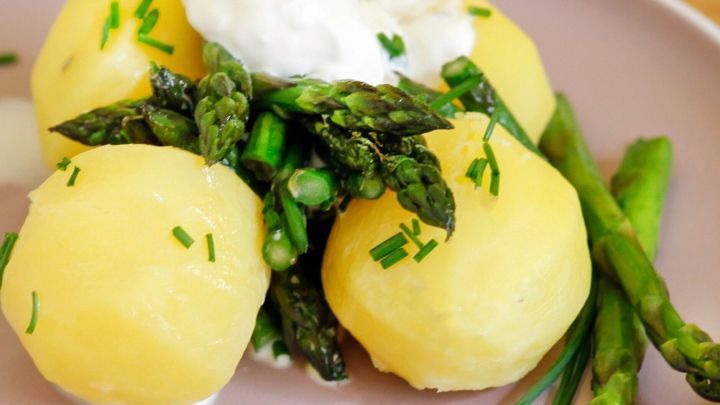 Gr&uuml;ner Spargel und Minikartoffeln liegen auf einem Teller, garniert mit Kr&auml;utern und einem Klecks Sahne.