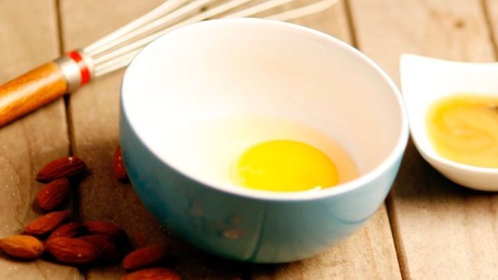 In einer Sch&uuml;ssel liegt ein rohes Eigelb, w&auml;hrend Mandeln und ein Schneebesen auf einem Holztisch liegen, bereit f&uuml;r das Mandel-Peeling.