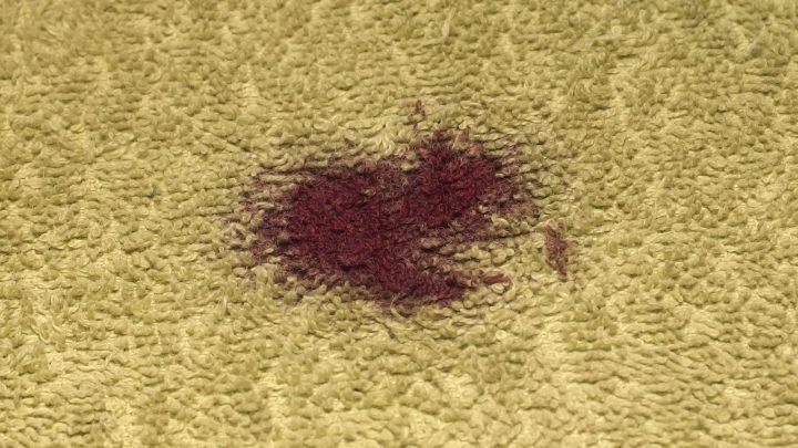 Ein Rotweinfleck liegt auf einem hellen Teppich; jemand könnte versuchen, ihn mit Glasreiniger zu entfernen.