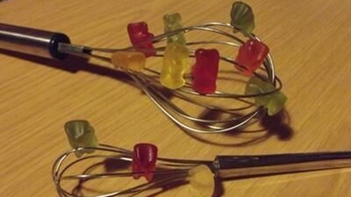 Bunte Gummib&auml;rchen sitzen auf einem Schneebesen, der auf einem Holzuntergrund liegt, als kreatives und verspieltes Arrangement.