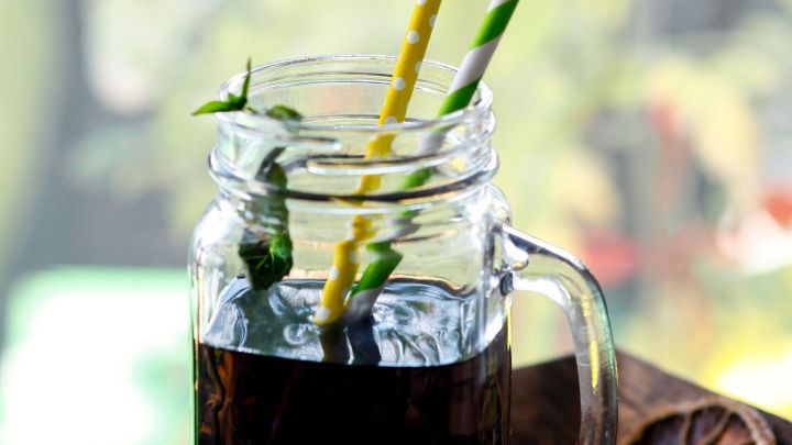 Ein Glas mit dunklem Getr&auml;nk steht auf einem Tisch, w&auml;hrend gelbe und gr&uuml;ne Strohhalme f&uuml;r Frische sorgen.