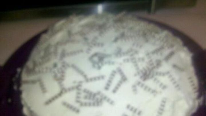 Ein Kuchen mit einer wei&szlig;en Zuckerglasur und bunten Streuseln liegt auf einem lila Teller in einer K&uuml;che.