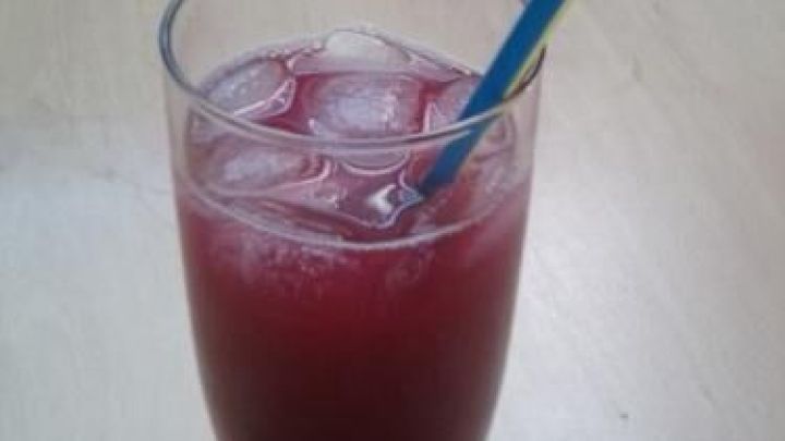 Ein Glas mit rotem Rhabarber-Eistee steht auf einem Holztisch, ein bunter Strohhalm ragt heraus.