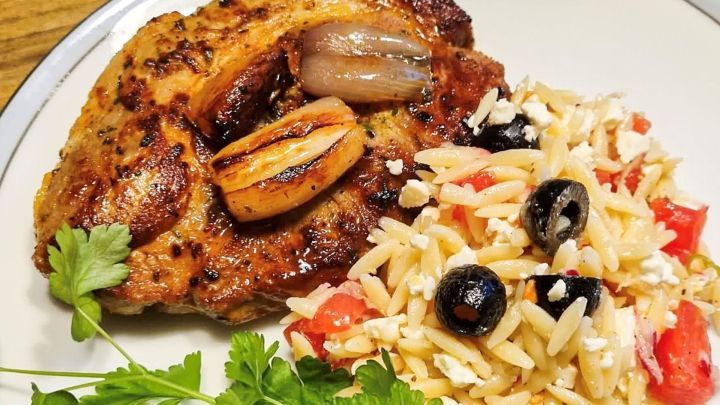 Saftiges, gegrilltes Fleischst&uuml;ck mit Zwiebeln liegt neben einer bunten Orzo-Salatbeilage auf einem Teller.