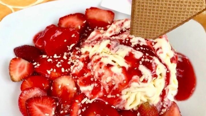 Ein Teller mit Spaghettieis zeigt eine Mischung aus Erdbeeren und Sahne, umgeben von roter Erdbeersauce und einem Waffelherz.