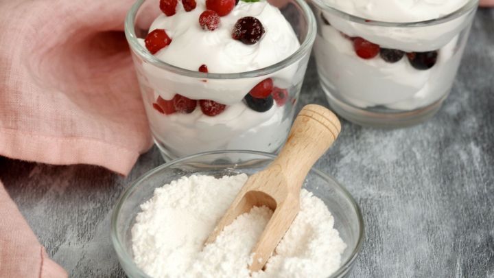 Zwei Gl&auml;ser mit Sahne und Fr&uuml;chten stehen neben einer Sch&uuml;ssel mit Zucker und einem kleinen L&ouml;ffel auf einem Tisch.