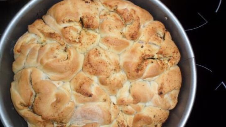 Faltenbrot mit einer goldbraunen Kruste liegt frisch gebacken in einer runden Backform auf einem dunklen Herd.